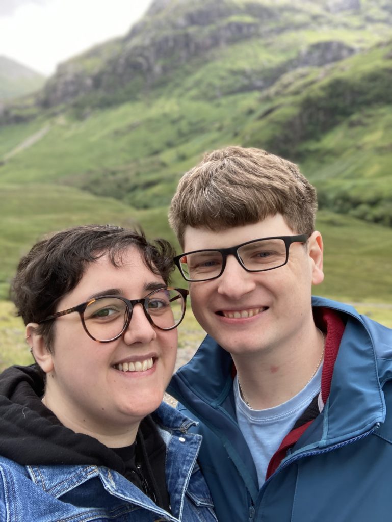 A selfie photo of James and Anna Sheasby Thomas with a hillside in the background. Anna is wearing a hoodie and a denim jacket, while James is wearing a raincoat, fleece and t shirt.